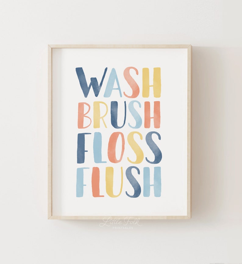 May include: A framed print with the words "WASH BRUSH FLOSS FLUSH" in colorful, watercolor-style lettering. The words are stacked vertically and are in shades of blue, yellow, and orange. The print is framed in a light wood frame.
