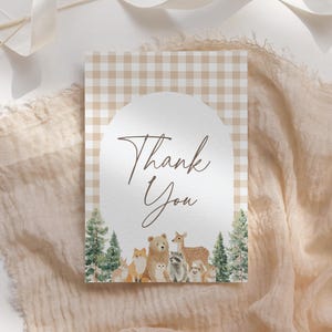 Peut inclure: Une carte de remerciement avec un fond à carreaux beiges et une arche blanche. La carte présente les mots "Thank You" dans une police marron. En dessous, une illustration d'animaux et d'arbres de la forêt.