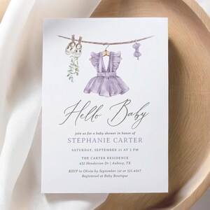 May include: A baby shower invitation with the words "Hello Baby" in elegant script. The invitation features a watercolor illustration of baby clothes hanging on a clothesline, including a purple dress. The details include the honoree's name, date, time, and location.
