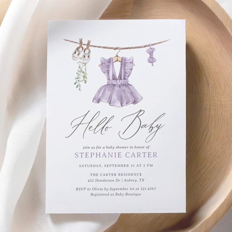 May include: A baby shower invitation with the words "Hello Baby" in elegant script. The invitation features a watercolor illustration of baby clothes hanging on a clothesline, including a purple dress. The details include the honoree's name, date, time, and location.