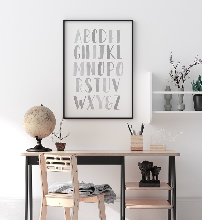 May include: A black framed poster with a white background and a silver alphabet printed on it. The poster is hanging on a white wall above a wooden desk with a globe, a potted plant, a pencil holder, and a wooden shelf with a small elephant figurine.