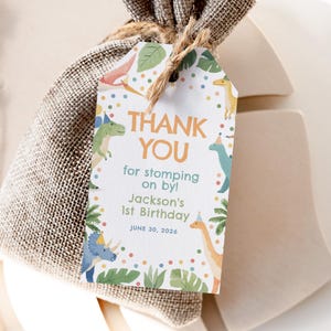 以下が含まれることがあります： 「THANK YOU」と「for stomping on by!」の文字、名前「Jackson's 1st Birthday」、日付「JUNE 30, 2026」が書かれた恐竜をテーマにしたギフトタグ。カラフルな恐竜のイラストと熱帯の葉で飾られています。
