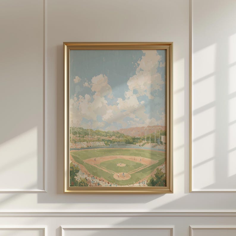 Op de afbeelding: Een ingelijst schilderij van een honkbalveld onder een bewolkte hemel. Het kunstwerk toont een groen veld, een bruin infield en een stadion vol toeschouwers. Het schilderij zit in een gouden lijst en is op een witte muur gemonteerd.