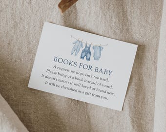 May include: A white card with the text "BOOKS FOR BABY" and a request for books instead of cards. The card features watercolor illustrations of baby clothes hanging on a clothesline. The card is on a neutral linen surface.
