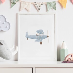 K&ouml;nnte beinhalten: Ein gerahmter Druck eines blauen Aquarell-Flugzeugs mit braunem Propeller und R&auml;dern. Der Druck ist auf wei&szlig;em Hintergrund und in einem wei&szlig;en Rahmen gerahmt.