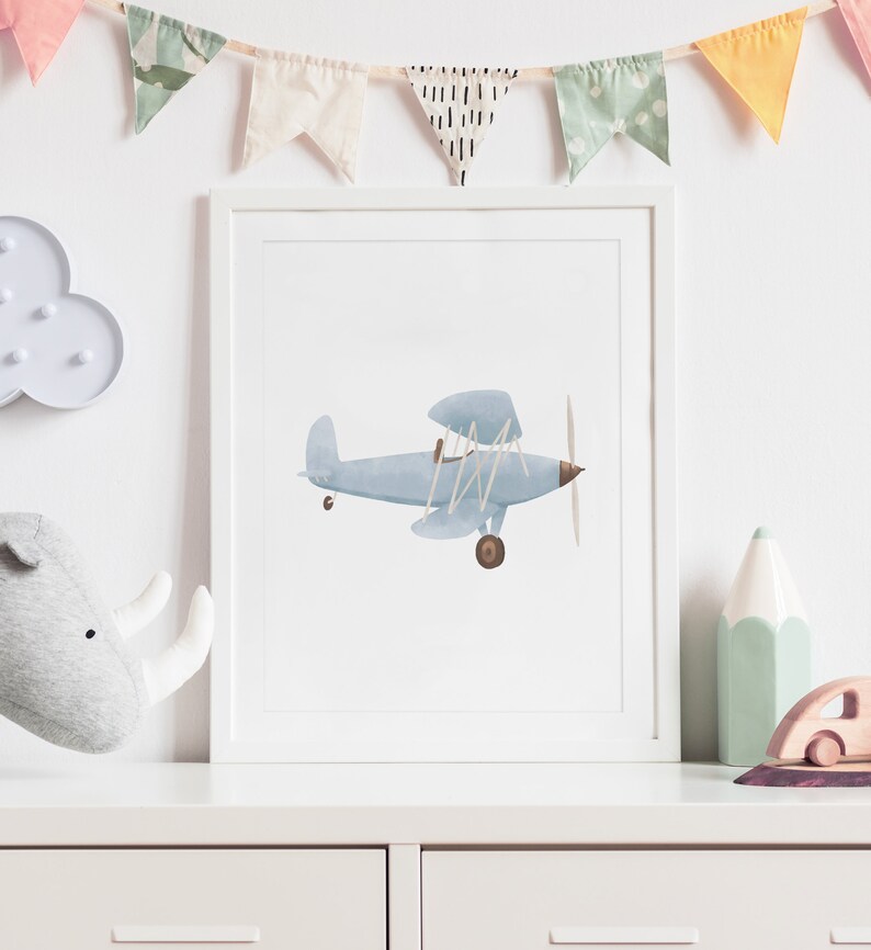 K&ouml;nnte beinhalten: Ein gerahmter Druck eines blauen Aquarell-Flugzeugs mit braunem Propeller und R&auml;dern. Der Druck ist auf wei&szlig;em Hintergrund und in einem wei&szlig;en Rahmen gerahmt.