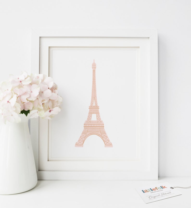 May include: A printable art print featuring a pink outline of the Eiffel Tower on a white background. The print is framed in a white frame and is sitting on a white surface. The print is from the company "littlefolk printables" and says "Original Artwork" at the bottom.