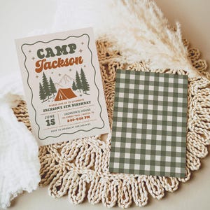 May include: A rectangular birthday invitation card featuring the text CAMP Jackson, a tent, pine trees, and mountain graphics in green and orange, alongside a second rectangular card with a green and white gingham check pattern.