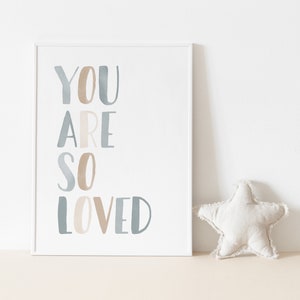 Puede incluir: Una impresi&oacute;n enmarcada en blanco con el texto "YOU ARE SO LOVED" en letras de acuarela azul y marr&oacute;n. Una almohada blanca en forma de estrella se encuentra en primer plano.