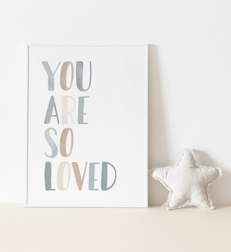 Puede incluir: Una impresi&oacute;n enmarcada en blanco con el texto "YOU ARE SO LOVED" en letras de acuarela azul y marr&oacute;n. Una almohada blanca en forma de estrella se encuentra en primer plano.