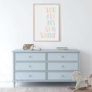 May include: A light blue dresser with six drawers and a wooden frame. The dresser has a light wood top and is decorated with a framed print that says "You Are My Sunshine" in colorful letters.