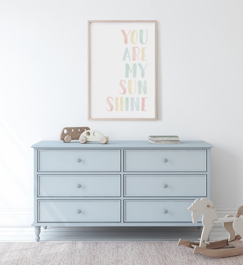 May include: A light blue dresser with six drawers and a wooden frame. The dresser has a light wood top and is decorated with a framed print that says "You Are My Sunshine" in colorful letters.