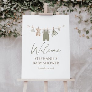 K&ouml;nnte beinhalten: Ein wei&szlig;es Willkommensschild f&uuml;r eine Babyparty auf einer Staffelei. Das Schild zeigt eine W&auml;scheleine mit Babykleidung und einer Teddyb&auml;r-Illustration. Der Text lautet "Welcome STEPHANIE'S BABY SHOWER September 21, 2026."