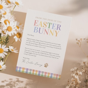 May include: A white card with the text "FROM THE DESK OF THE EASTER BUNNY" in pastel colors. The card includes a message to "Macey" and is decorated with a paw print. Daisies and other flowers are in the background.