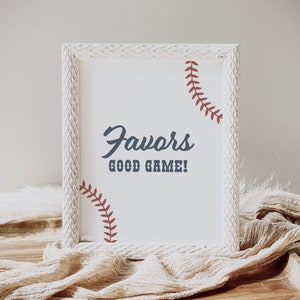 May include: White wooden frame with a white background and the text "Favors Good Game!" in blue. The frame has a baseball stitch design in red on the top and bottom.
