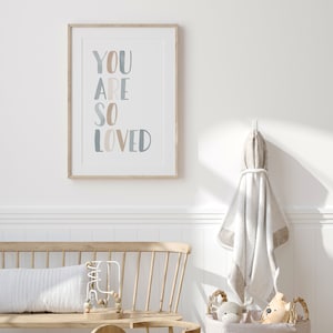 Puede incluir: Una impresi&oacute;n enmarcada con las palabras "YOU ARE SO LOVED" en un estilo acuarela azul claro y beige. La impresi&oacute;n est&aacute; colgada en una pared blanca sobre un banco de madera con un coj&iacute;n blanco y un juguete de madera.