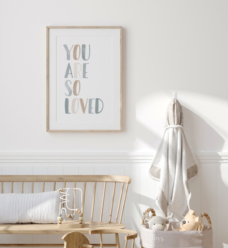 Puede incluir: Una impresi&oacute;n enmarcada con las palabras "YOU ARE SO LOVED" en un estilo acuarela azul claro y beige. La impresi&oacute;n est&aacute; colgada en una pared blanca sobre un banco de madera con un coj&iacute;n blanco y un juguete de madera.