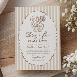 Può includere: Invito per baby shower con sfondo a righe beige e bianco. L'invito presenta una cornice ovale marrone con un'illustrazione di un cestino di pane e il testo "There's a Bun in the Oven". Ulteriori dettagli sono inclusi.