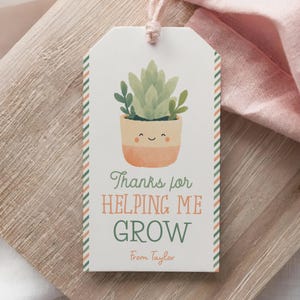 May include: A white gift tag with a watercolor illustration of a potted succulent plant. The tag has a striped border and reads "Thanks for Helping Me Grow" with a name below. A pink string is attached.