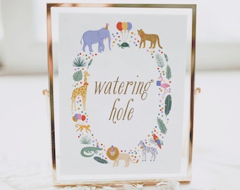 May include: A framed print with a gold-colored metal frame. The print features the words "watering hole" in gold script, surrounded by colorful illustrations of animals like an elephant, giraffe, and lion. The background is white.