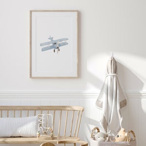 May include: A framed watercolor illustration of a blue airplane against a white background. The airplane is a biplane with a propeller. The frame is light wood.