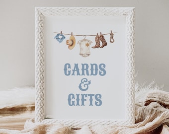 Op de afbeelding: Een wit ingelijst bord met een blauwe achtergrond en de tekst "CARDS & GIFTS". Het bord is versierd met een waslijn met een blauw shirt, een cowboyhoed, een baby-romper en cowboylaarzen die eraan hangen.