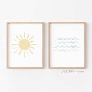 Puede incluir: Dos láminas de arte mural enmarcadas en madera clara. La lámina de la izquierda muestra una ilustración de un sol amarillo sobre fondo blanco. La de la derecha muestra líneas onduladas azul claro que representan agua. El texto Little Folk Printables es visible.