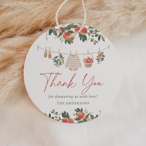 May include: Round white tag with a string, featuring a baby shower theme. The tag has a watercolor design with apples, greenery, and baby clothes. Text reads "Thank You" in red script, with additional text below.