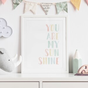 May include: A white framed print with the text "YOU ARE MY SUN SHINE" in rainbow colors. The print is hanging on a white wall with a string of colorful flags above it.