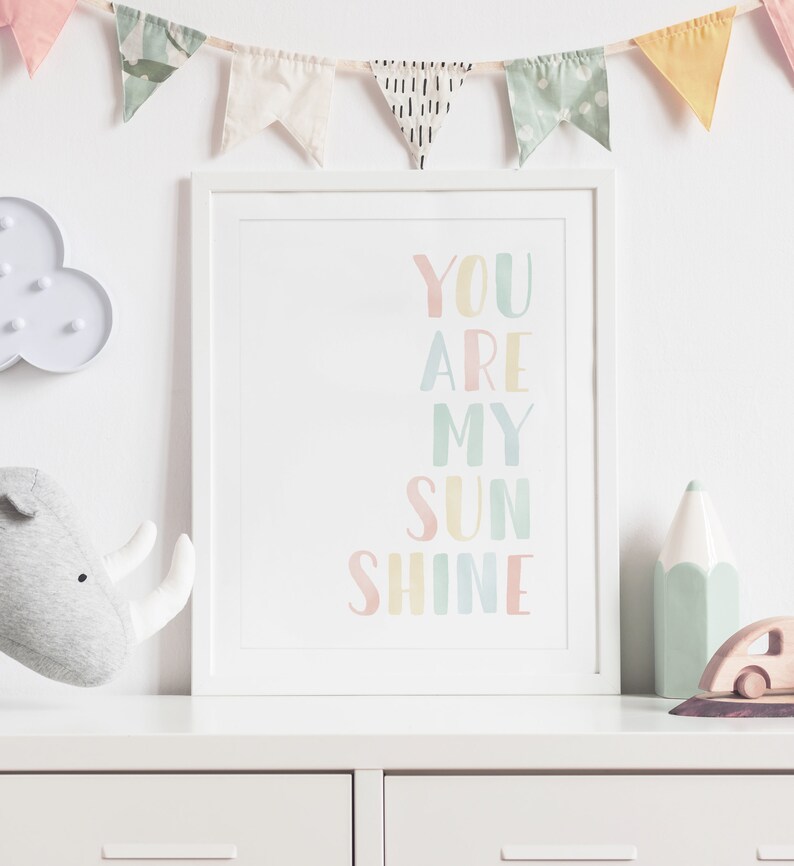 May include: A white framed print with the text "YOU ARE MY SUN SHINE" in rainbow colors. The print is hanging on a white wall with a string of colorful flags above it.