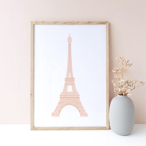 May include: A framed print of a pink outline of the Eiffel Tower on a white background. The frame is light wood and the print is sitting on a white surface.