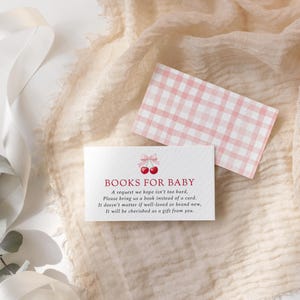 May include: A baby shower invitation with the text "BOOKS FOR BABY" and a cherry graphic. The invitation is white with red text and a pink gingham pattern card. The invitation requests a book instead of a card.