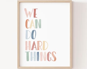Puede incluir: Una lámina artística enmarcada con el texto WE CAN DO HARD THINGS en un estilo de acuarela multicolor. Las letras están representadas en tonos rosa, naranja, azul y verde. El marco es de madera clara y el texto Little Folk Printables aparece en la parte inferior.