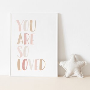 K&ouml;nnte beinhalten: Ein wei&szlig;er gerahmter Druck mit dem Text "YOU ARE SO LOVED" in rosa und braunen Aquarellbuchstaben.