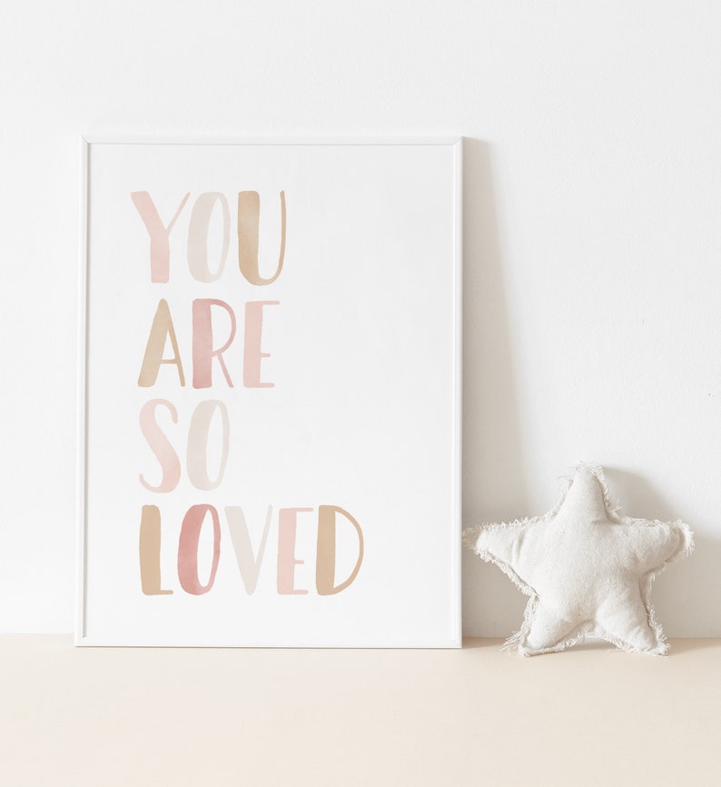K&ouml;nnte beinhalten: Ein wei&szlig;er gerahmter Druck mit dem Text "YOU ARE SO LOVED" in rosa und braunen Aquarellbuchstaben.