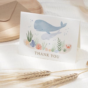 May include: A watercolor thank you card with a blue whale and a baby whale swimming in the ocean. The card features coral, seaweed, and a starfish. The text "THANK YOU" is printed in gold on the card.