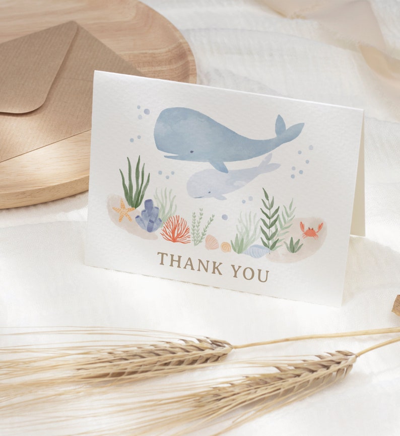 May include: A watercolor thank you card with a blue whale and a baby whale swimming in the ocean. The card features coral, seaweed, and a starfish. The text "THANK YOU" is printed in gold on the card.