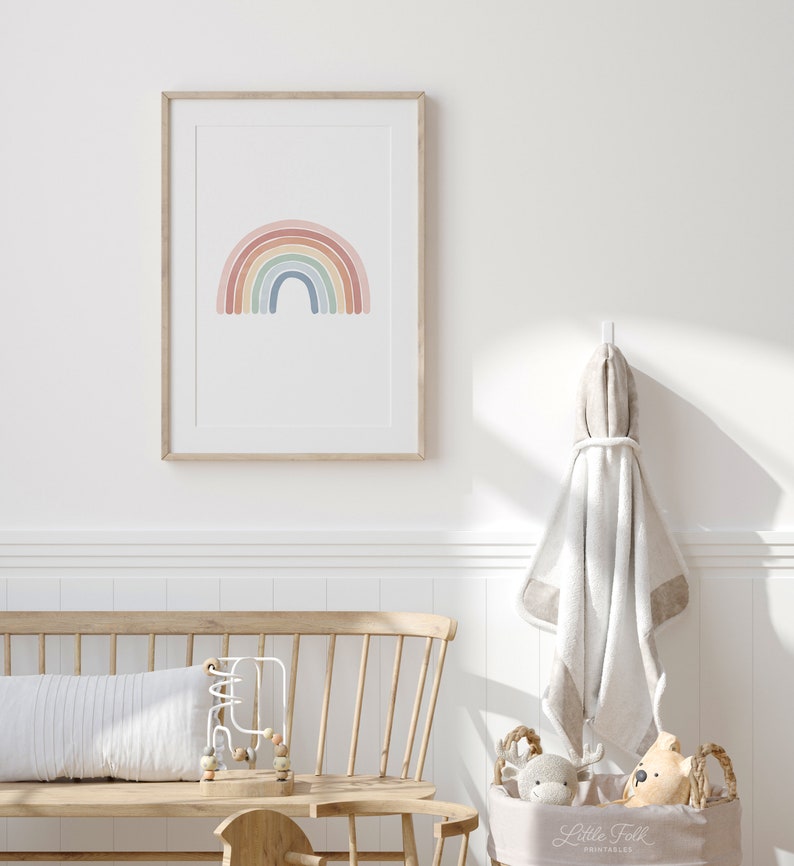 K&ouml;nnte beinhalten: Ein gerahmter Druck eines Regenbogens mit Pastellfarben in einem Kinderzimmer. Der Regenbogen befindet sich in einem Holzrahmen und h&auml;ngt an einer wei&szlig;en Wand. Im Vordergrund befindet sich eine Holzbank mit einem wei&szlig;en Kissen und einem Korb mit Stofftieren.