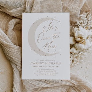 May include: A cream-colored invitation card with the text "She's Over the Moon" in elegant script, featuring a crescent moon and star design. The card includes event details: Cassidy Michaels' bridal shower, date, time, and location. The invitation is set against a neutral fabric.