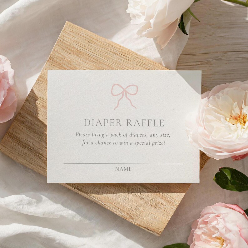 Może przedstawiać: Prostokątna karta z napisem "DIAPER RAFFLE" i r&oacute;żową ilustracją kokardy. Karta leży na drewnianej podstawie, otoczona r&oacute;żowymi kwiatami. Tekst zaprasza do przyniesienia pieluch, aby wygrać nagrodę.