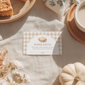 May include: A diaper raffle card with a tan and white gingham pattern. The card features the text "DIAPER RAFFLE" and instructions. A small pie graphic is at the top. The card is on a neutral-toned surface.