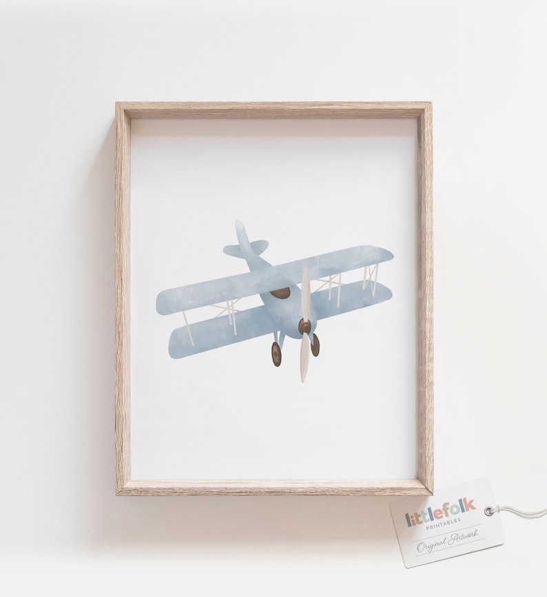 May include: A framed print of a blue watercolor illustration of a vintage airplane with a propeller. The print is in a light wood frame and has a white background. The text "littlefolk printables Original Artwork" is on a tag in the bottom right corner of the image.