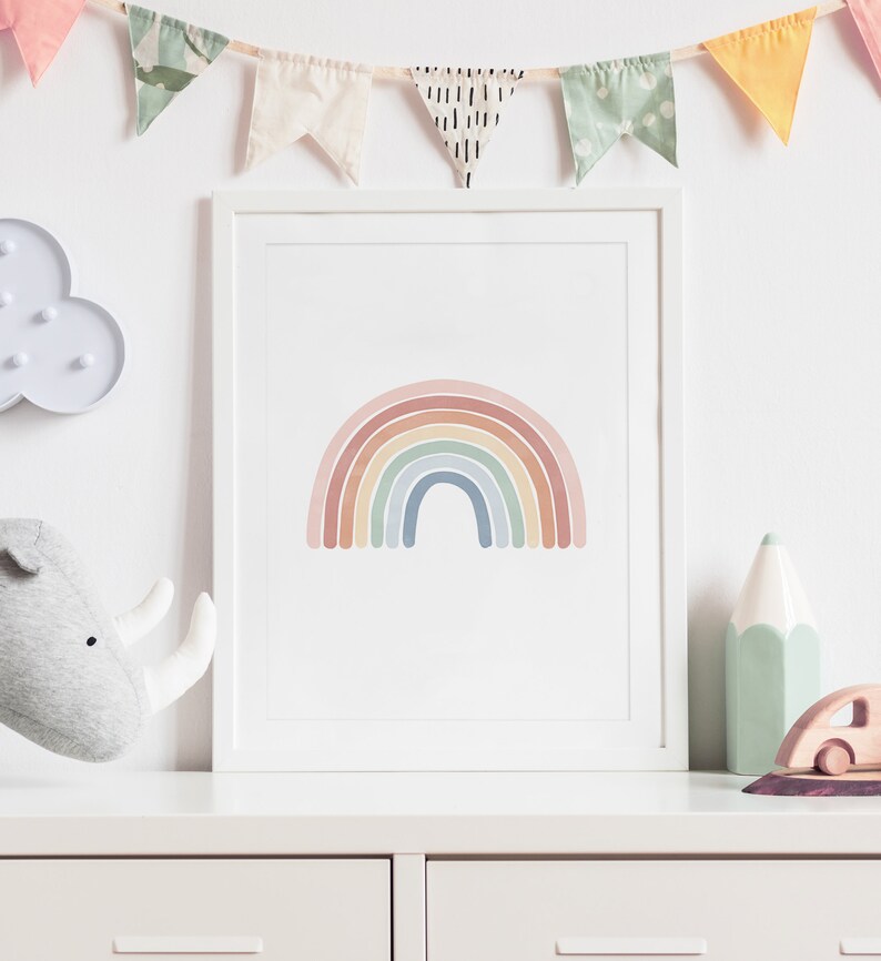 K&ouml;nnte beinhalten: Ein gerahmter Druck eines Regenbogens in Pastellfarben, der an einer wei&szlig;en Wand &uuml;ber einer wei&szlig;en Kommode mit einem Spielzeugnashorn, einem gr&uuml;nen Bleistift und einem Holzspielzeugauto h&auml;ngt.