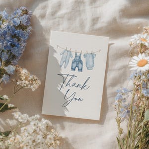 May include: A baby shower thank you card with watercolor illustrations of baby clothes hanging on a clothesline. The card features a blue and white color scheme with the words "Thank You" in elegant script.