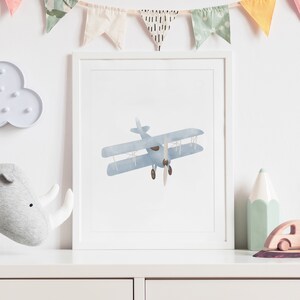 May include: A framed print of a blue watercolor airplane with brown propeller blades. The print is in a white frame and is hanging on a white wall.
