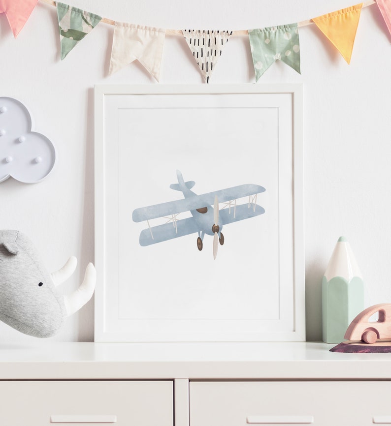 May include: A framed print of a blue watercolor airplane with brown propeller blades. The print is in a white frame and is hanging on a white wall.