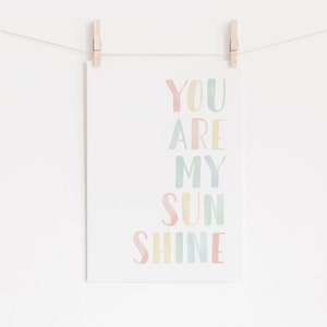 May include: A pastel-colored print with the text "You are my sunshine" in a rainbow of colors. The print is hanging on a white wall with clothespins.
