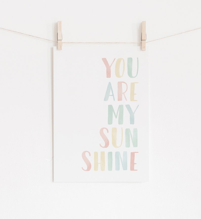 May include: A pastel-colored print with the text "You are my sunshine" in a rainbow of colors. The print is hanging on a white wall with clothespins.