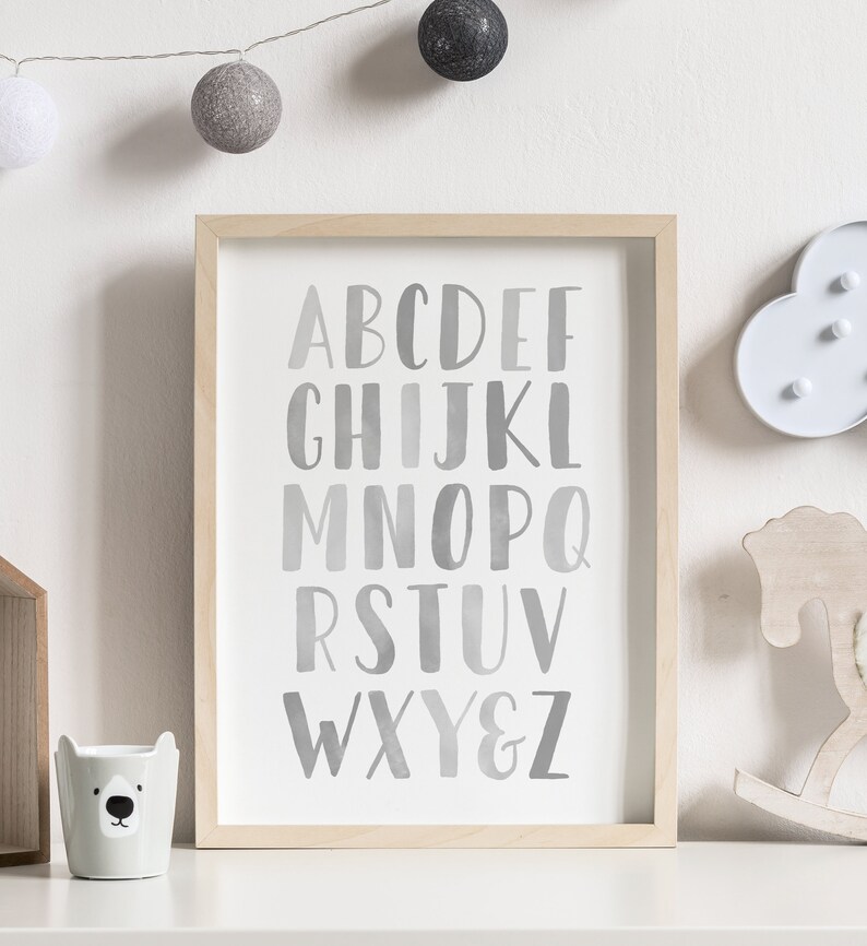 May include: A framed print with a white background and a gray watercolor alphabet. The letters are in a gradient of gray, starting with a light gray and fading to a darker gray. The letters are arranged in three rows, with the first row being A-F, the second row being G-L, and the third row being M-Z. The frame is a light wood color.