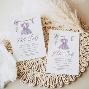 May include: Two baby shower invitations with a watercolor design. Each card features a purple dress hanging on a clothesline, with the words "Hello Baby" in elegant script. The invitations are on a white background.